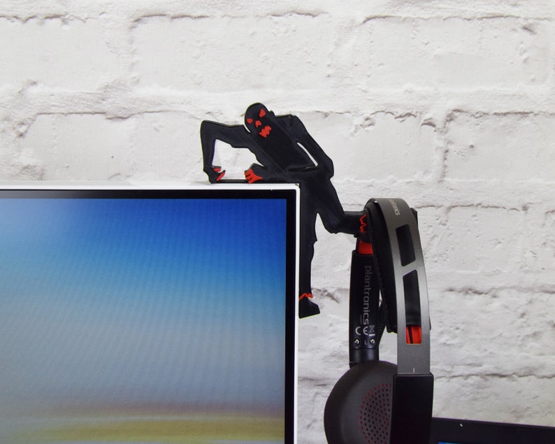 May include: A black 3D printed figure of a monster with red eyes is holding a pair of black headphones with red accents. The headphones have the text "Plantronics" on the side. The figure is perched on the top of a black computer monitor.