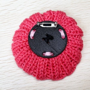 May include: A pink knitted flower brooch with a black button and a silver pin on the back.