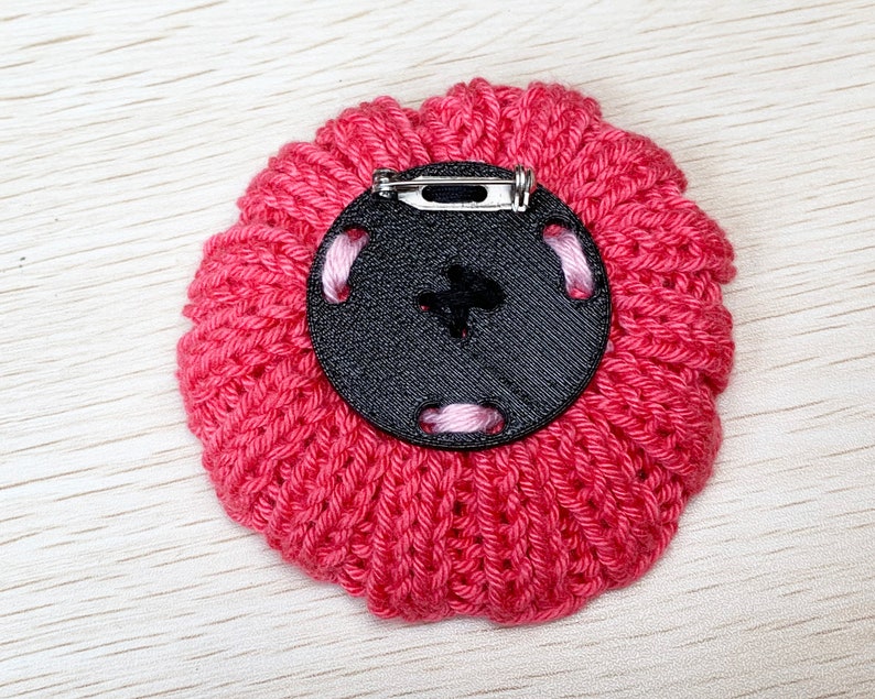 May include: A pink knitted flower brooch with a black button and a silver pin on the back.