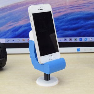 May include: A blue and white 3D printed phone stand shaped like a hand holding a phone. The phone stand is on a wooden surface.