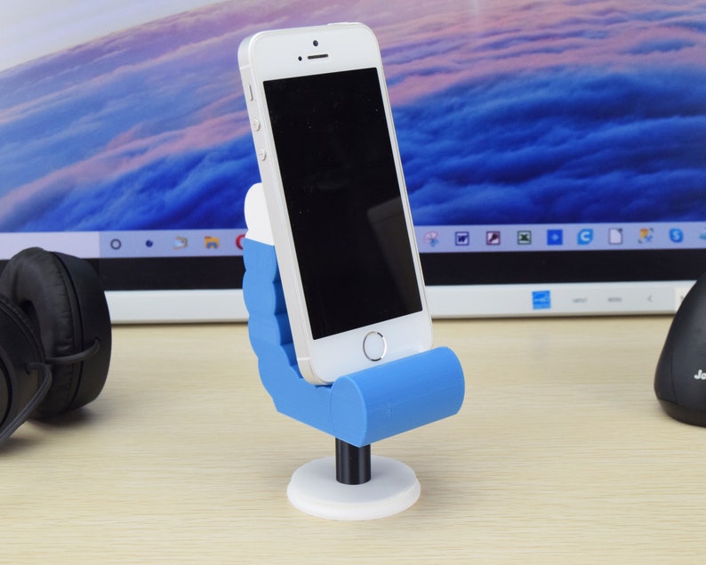 May include: A blue and white 3D printed phone stand shaped like a hand holding a phone. The phone stand is on a wooden surface.
