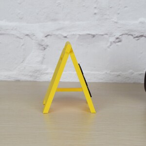May include: A yellow plastic construction sign shaped like a triangle with a black base. The sign is standing on a light brown wooden surface.