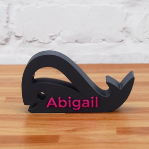 May include: Black whale-shaped name holder with the name "Abigail" printed in pink.