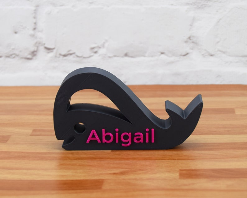May include: Black whale-shaped name holder with the name "Abigail" printed in pink.
