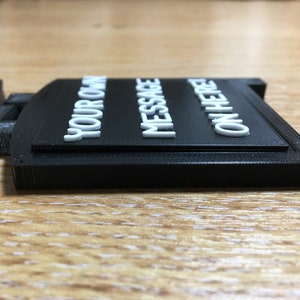 May include: A black rectangular sign with the words "YOUR OWN MESSAGE ON HERE" in white, raised lettering. The sign is made of a matte material and sits on a wooden surface.