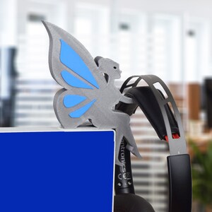 K&ouml;nnte beinhalten: Eine graue, 3D-gedruckte Schmetterlingsfigur mit blauen Fl&uuml;geln sitzt auf einer blauen Oberfl&auml;che. Hinter der Figur befindet sich ein schwarzes und rotes Headset.