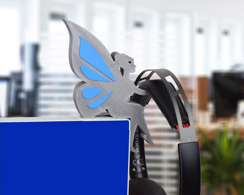 K&ouml;nnte beinhalten: Eine graue, 3D-gedruckte Schmetterlingsfigur mit blauen Fl&uuml;geln sitzt auf einer blauen Oberfl&auml;che. Hinter der Figur befindet sich ein schwarzes und rotes Headset.