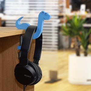 May include: Black headphones with a blue plastic snake-shaped headphone holder. The headphones are hanging on a wooden shelf.