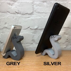 May include: Two phone stands made of 3D printed plastic in the shape of a cartoon worm. The stand on the left is gray and holds a smartphone. The stand on the right is silver and holds a tablet.