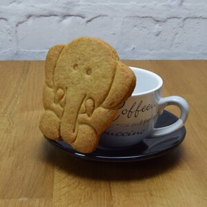 May include: A light brown elephant-shaped cookie sits on a saucer next to a white coffee mug with the words "Coffee, rice and fruit, cappuccino" printed on it.