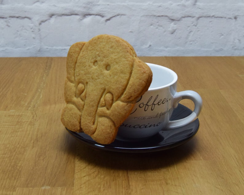 May include: A light brown elephant-shaped cookie sits on a saucer next to a white coffee mug with the words "Coffee, rice and fruit, cappuccino" printed on it.