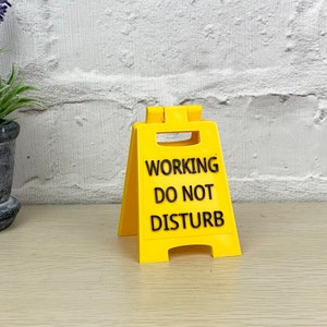 Op de afbeelding: Een geel plastic bord met zwarte tekst die "WORKING DO NOT DISTURB" luidt.