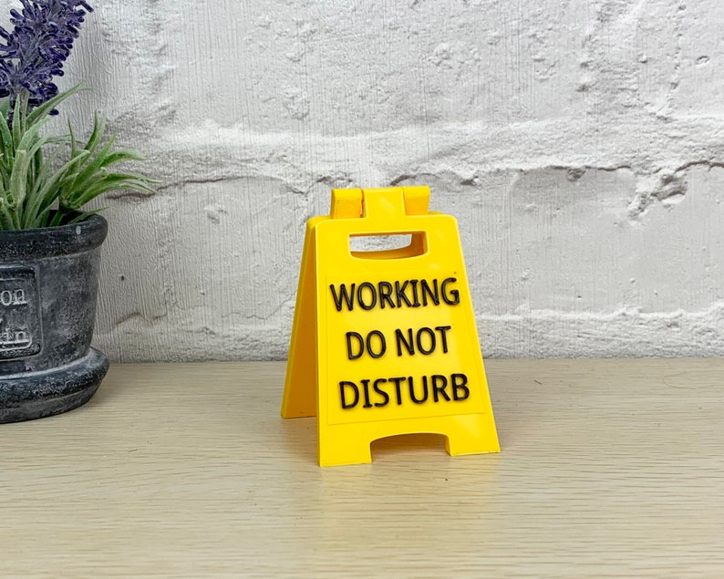 Op de afbeelding: Een geel plastic bord met zwarte tekst die "WORKING DO NOT DISTURB" luidt.