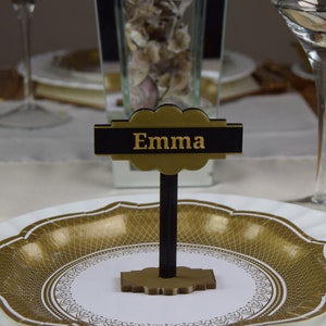 Może przedstawiać: Złota i czarna karta stołowa z napisem "Emma" w złotych literach. Karta stoi na czarnym stojaku i leży na białym talerzu z złotym wzorem.