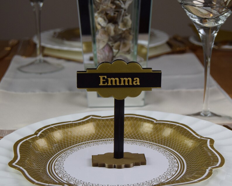 Może przedstawiać: Złota i czarna karta stołowa z napisem "Emma" w złotych literach. Karta stoi na czarnym stojaku i leży na białym talerzu z złotym wzorem.