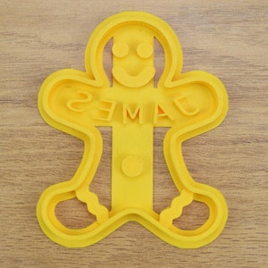 Puede incluir: Cortador de galletas de jengibre de pl&aacute;stico amarillo en forma de hombre con el nombre "JAMES" escrito en el medio.