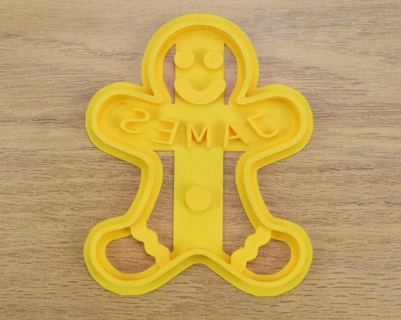 Puede incluir: Cortador de galletas de jengibre de pl&aacute;stico amarillo en forma de hombre con el nombre "JAMES" escrito en el medio.