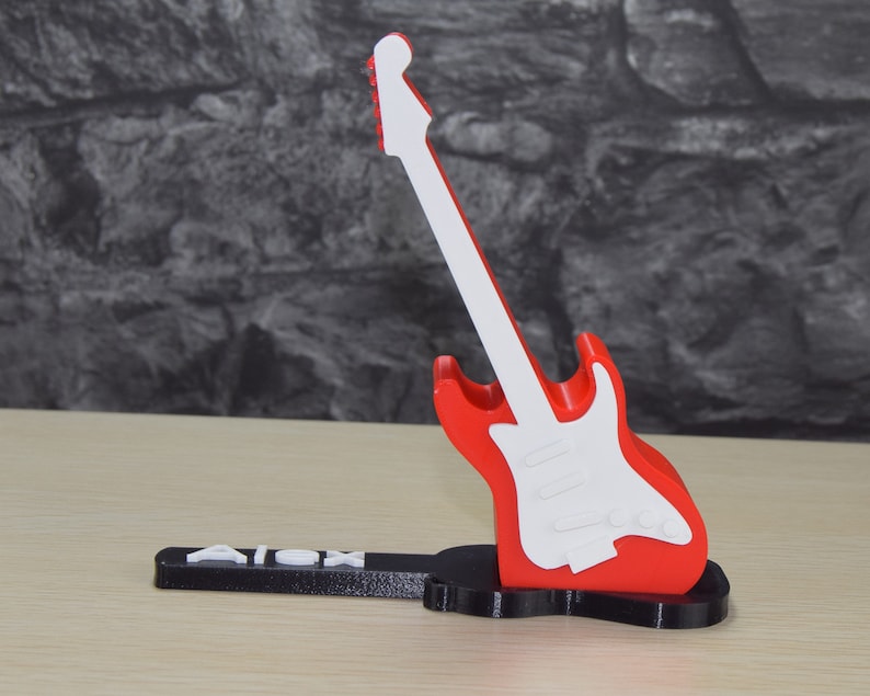 Puede incluir: Un soporte para tel&eacute;fono de guitarra impreso en 3D, rojo y blanco, con el nombre "Alex" impreso en la base.