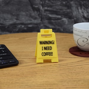 May include: A yellow warning sign with black text that reads "WARNING! I NEED COFFEE!" The sign is shaped like a traditional construction sign with a slanted top and two legs.