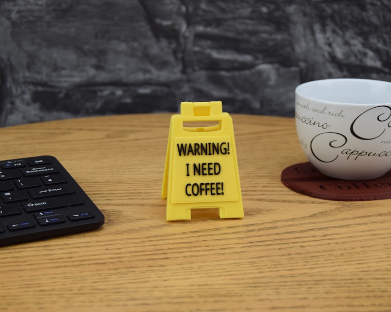 May include: A yellow warning sign with black text that reads "WARNING! I NEED COFFEE!" The sign is shaped like a traditional construction sign with a slanted top and two legs.