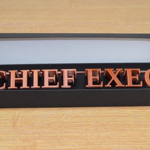 May include: A black box with the lid open, revealing a set of copper-colored letters spelling out "CHIEF EXEC" in a decorative font.