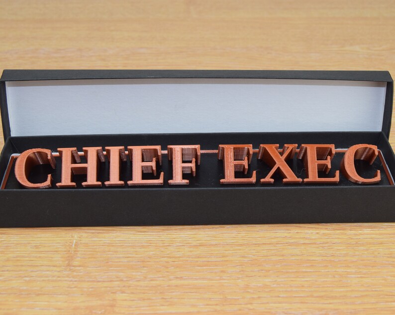 May include: A black box with the lid open, revealing a set of copper-colored letters spelling out "CHIEF EXEC" in a decorative font.
