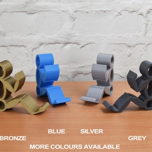 May include: Four phone stands made of plastic in the colors bronze, blue, silver, and grey. The stands are shaped like stick figures sitting down with their arms raised.
