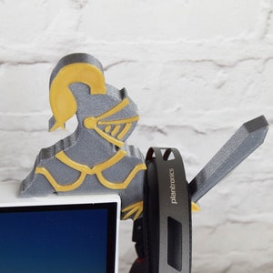 Op de afbeelding: Een 3D-geprint grijs en gouden ridderfiguur met een zwaard, dat een paar zwarte koptelefoons vasthoudt met het woord "Plantronics" erop gedrukt.