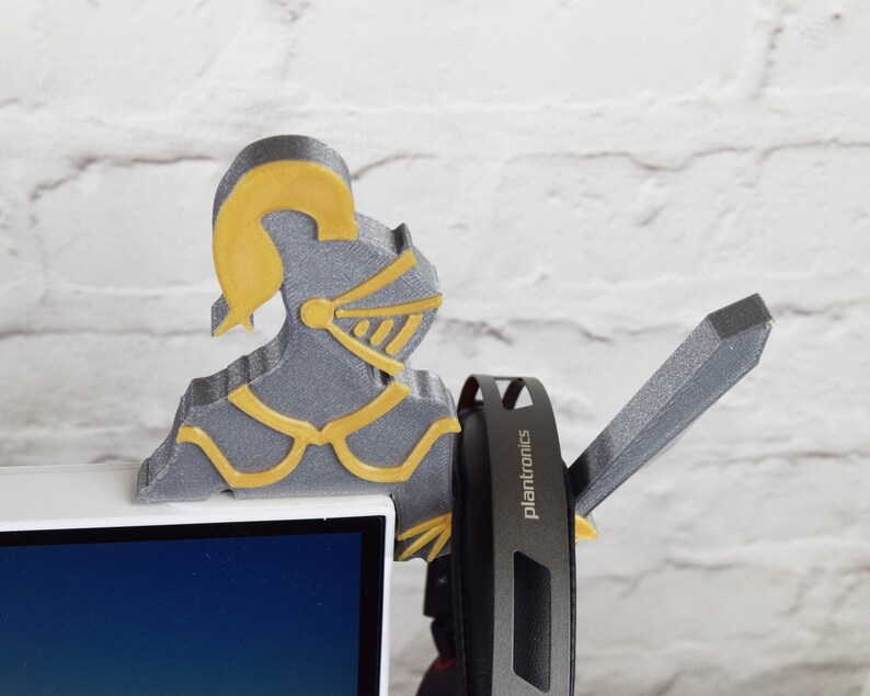 Op de afbeelding: Een 3D-geprint grijs en gouden ridderfiguur met een zwaard, dat een paar zwarte koptelefoons vasthoudt met het woord "Plantronics" erop gedrukt.