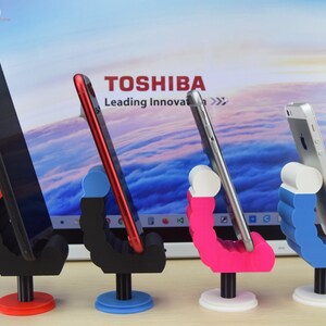 May include: Four phone stands with different colored bases. The stands are made of plastic and have a unique design that allows them to hold a phone in a vertical position. The stands are in front of a laptop screen with the Toshiba logo and the text "Leading Innovation".