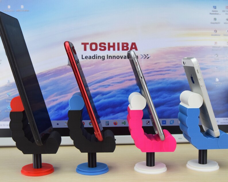 May include: Four phone stands with different colored bases. The stands are made of plastic and have a unique design that allows them to hold a phone in a vertical position. The stands are in front of a laptop screen with the Toshiba logo and the text "Leading Innovation".