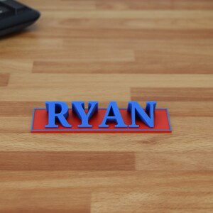 May include: A 3D-printed nameplate with the name "RYAN" in blue letters on a red rectangular base. The nameplate is set on a wooden surface, with a black keyboard visible in the background.