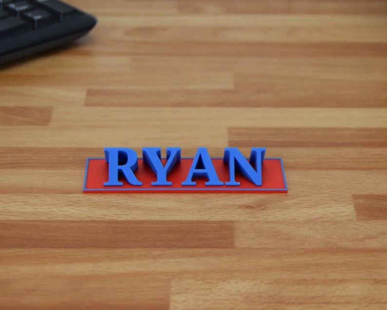 May include: A 3D-printed nameplate with the name "RYAN" in blue letters on a red rectangular base. The nameplate is set on a wooden surface, with a black keyboard visible in the background.