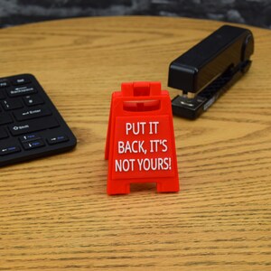 May include: A red plastic sign with the text "PUT IT BACK, IT'S NOT YOURS!" stands on a wooden desk. A black stapler and a black wireless keyboard are also on the desk.