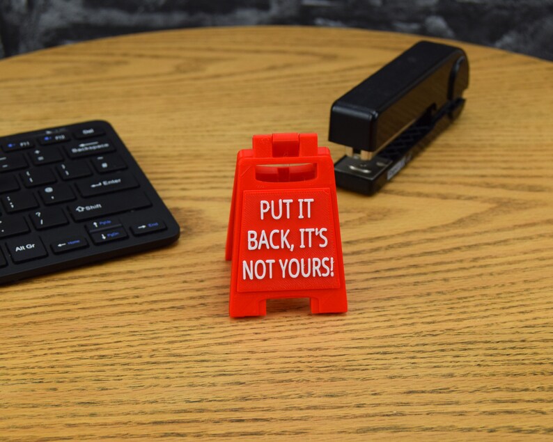 May include: A red plastic sign with the text "PUT IT BACK, IT'S NOT YOURS!" stands on a wooden desk. A black stapler and a black wireless keyboard are also on the desk.