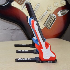 Puede incluir: Tres soportes para tel&eacute;fono impresos en 3D con forma de guitarras el&eacute;ctricas. Cada soporte tiene un nombre diferente en la base: Jack, Ted y Alex. Los soportes son rojos, blancos y azules.
