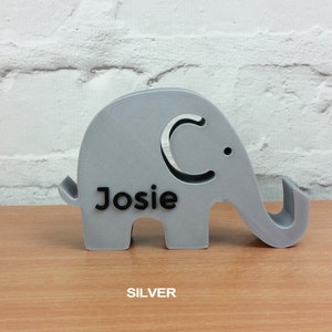 Pode incluir: Um suporte de telefone de elefante prateado com o nome "Josie" impresso nele.