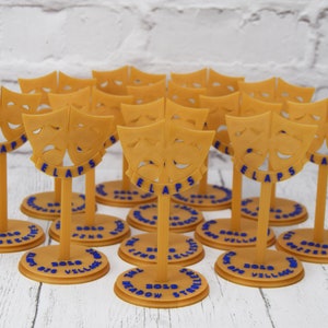 May include: A group of 14 gold-colored, 3D printed, theatrical mask award trophies. Each trophy features a pair of comedic and tragic masks with the word "FLAPS" printed below. The base of each trophy is a round, gold-colored platform with the words "2020 BOLD" and the name of a play printed on it.