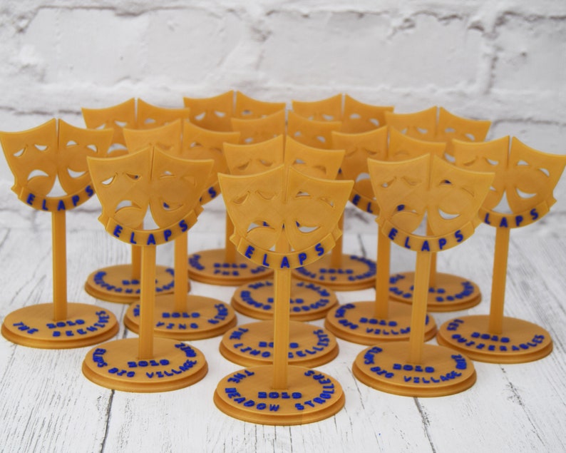 May include: A group of 14 gold-colored, 3D printed, theatrical mask award trophies. Each trophy features a pair of comedic and tragic masks with the word "FLAPS" printed below. The base of each trophy is a round, gold-colored platform with the words "2020 BOLD" and the name of a play printed on it.