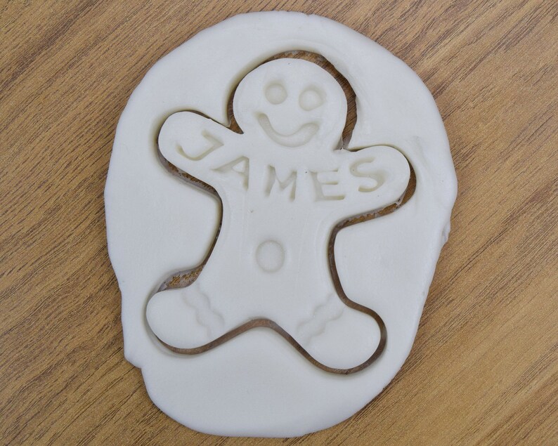 Puede incluir: Cortador de galletas de jengibre blanco con el nombre "JAMES" cortado en el centro.