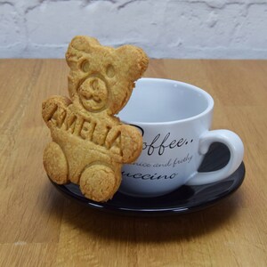 Puede incluir: Una galleta con forma de oso marr&oacute;n con el nombre "Amelia" en relieve, sentada en un platillo negro junto a una taza de caf&eacute; blanca con el texto "coffee... nice and frothyuccino" impreso en ella.