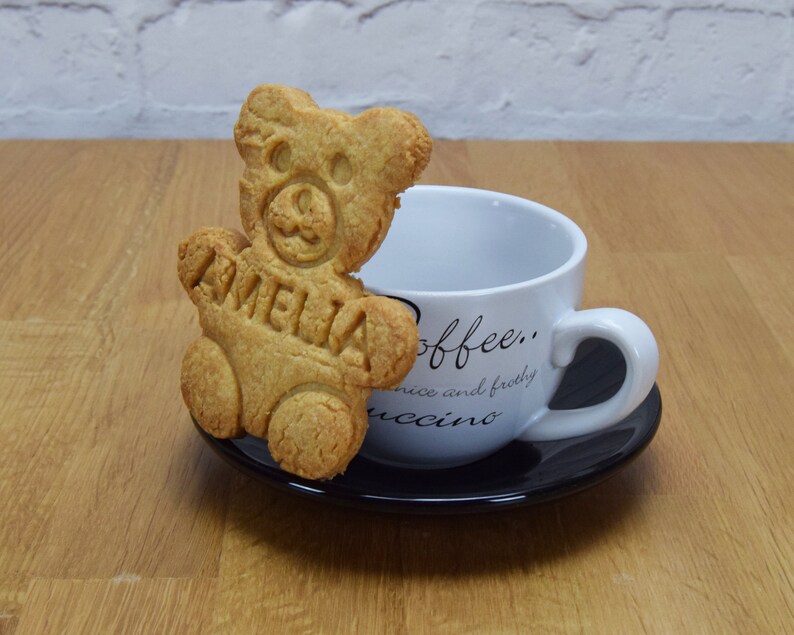 Puede incluir: Una galleta con forma de oso marr&oacute;n con el nombre "Amelia" en relieve, sentada en un platillo negro junto a una taza de caf&eacute; blanca con el texto "coffee... nice and frothyuccino" impreso en ella.