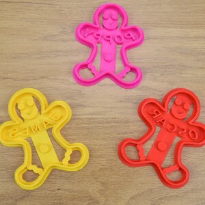 Puede incluir: Tres cortadores de galletas de hombre de jengibre, uno rojo, uno amarillo y uno rosa. Los cortadores tienen forma de hombre de jengibre con los brazos extendidos y tienen los nombres "Oscar", "James" y "Poppy" escritos en ellos.
