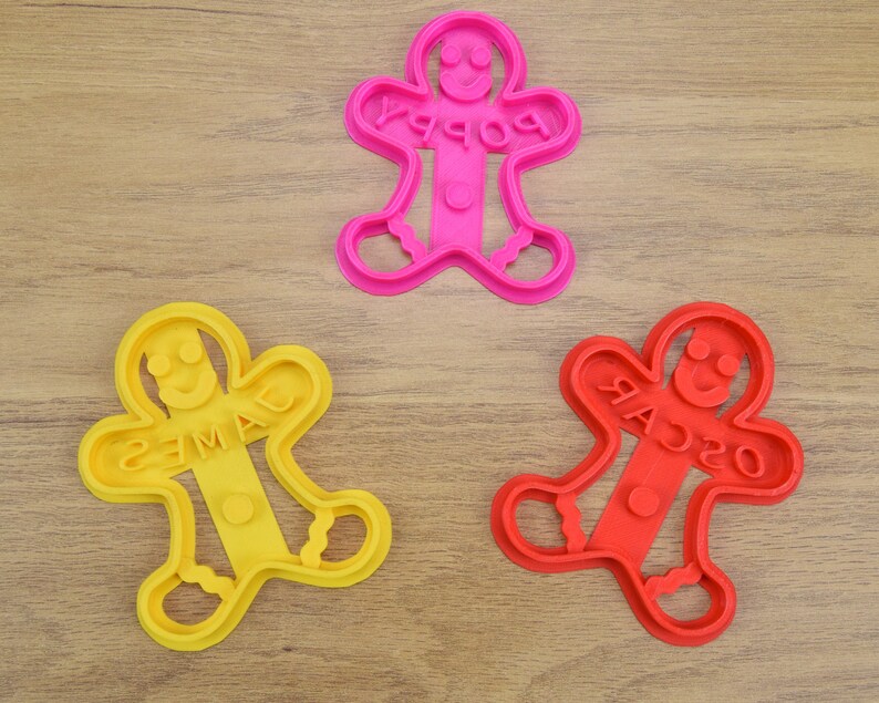 Puede incluir: Tres cortadores de galletas de hombre de jengibre, uno rojo, uno amarillo y uno rosa. Los cortadores tienen forma de hombre de jengibre con los brazos extendidos y tienen los nombres "Oscar", "James" y "Poppy" escritos en ellos.