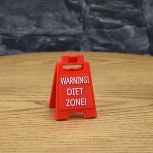 Może przedstawiać: Czerwony plastikowy znak ostrzegawczy z napisem "WARNING! DIET ZONE!" na drewnianej powierzchni.