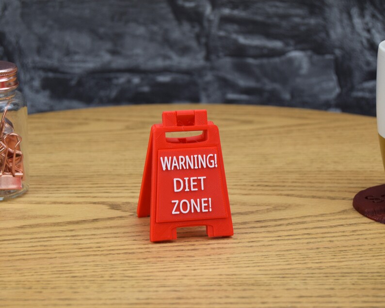 Może przedstawiać: Czerwony plastikowy znak ostrzegawczy z napisem "WARNING! DIET ZONE!" na drewnianej powierzchni.