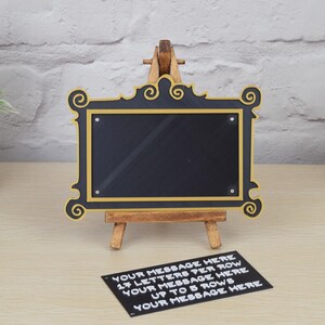 May include: A black chalkboard with a gold frame, resembling a television screen, sits on a wooden easel. The chalkboard is blank, but a note below it reads "YOUR MESSAGE HERE 17 LETTERS PER ROW YOUR MESSAGE HERE UP TO 5 ROWS YOUR MESSAGE HERE".