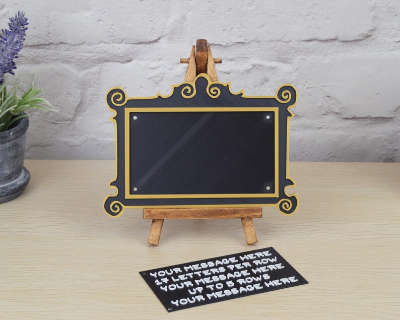 May include: A black chalkboard with a gold frame, resembling a television screen, sits on a wooden easel. The chalkboard is blank, but a note below it reads "YOUR MESSAGE HERE 17 LETTERS PER ROW YOUR MESSAGE HERE UP TO 5 ROWS YOUR MESSAGE HERE".