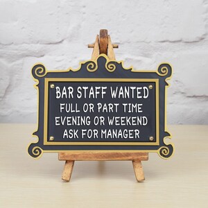 May include: Black sign with gold trim and white text on a wooden easel. The sign reads "Bar Staff Wanted Full or Part Time Evening or Weekend Ask for Manager".
