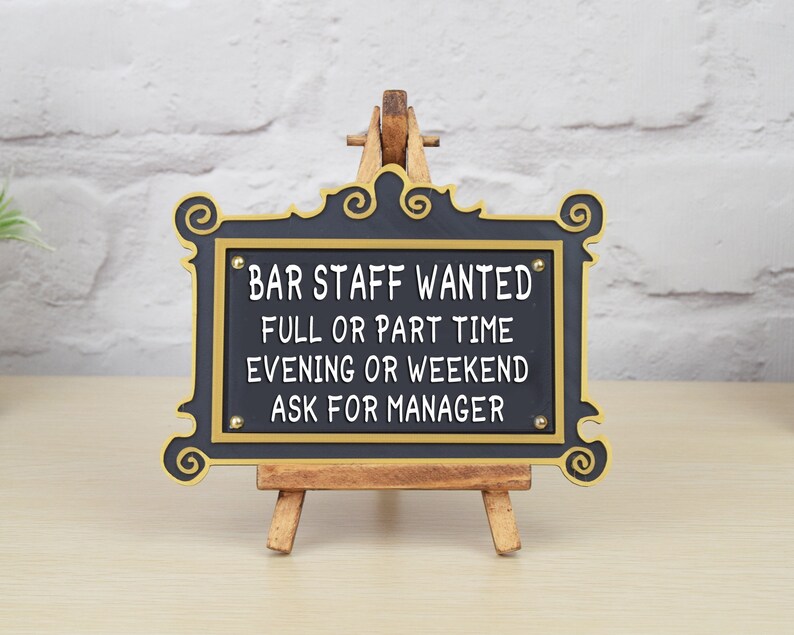 May include: Black sign with gold trim and white text on a wooden easel. The sign reads "Bar Staff Wanted Full or Part Time Evening or Weekend Ask for Manager".